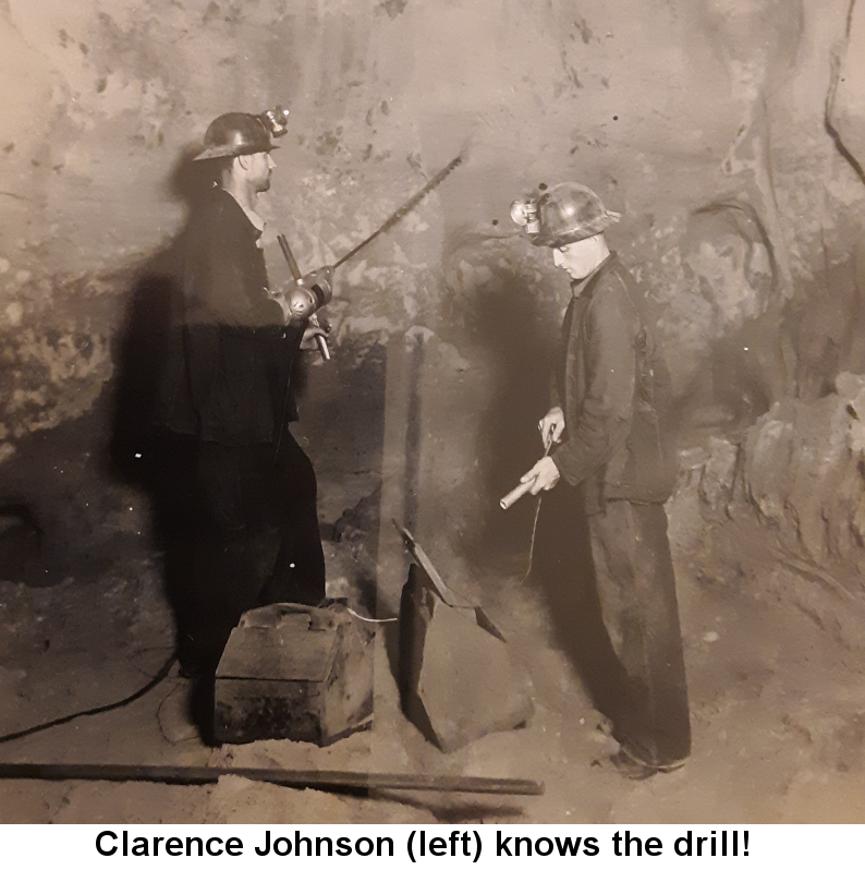 Colorized photo of Clarence Casper Johnson with helmet and dark work clothes, holding an electric drill with a very long bit near a rock wall in the sand mine. At his feet are a satchel of explosives and a wooden toolbox; next to him stands another miner inserting a fuse into a stick of dynamite.
